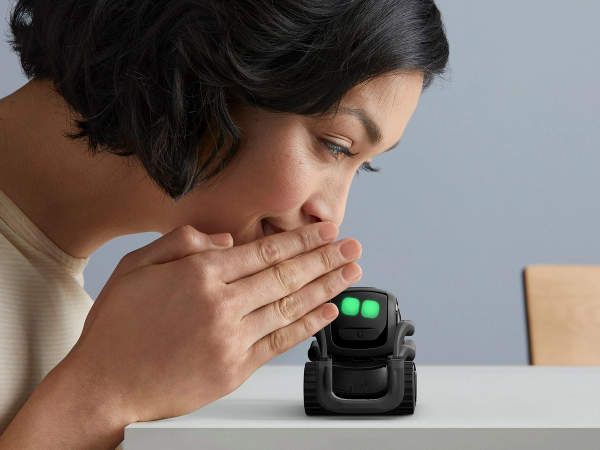A woman whispers to a small wheeled robot with large green eyes called Vector