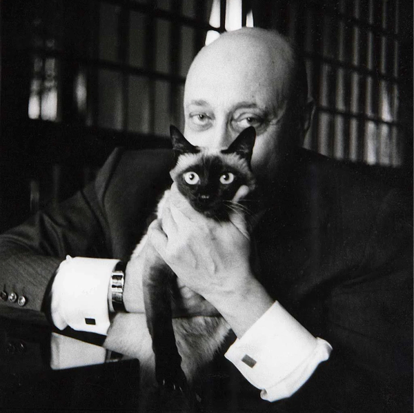 A black and white photo of a bald man obscuring his face by holding a Siamese cat