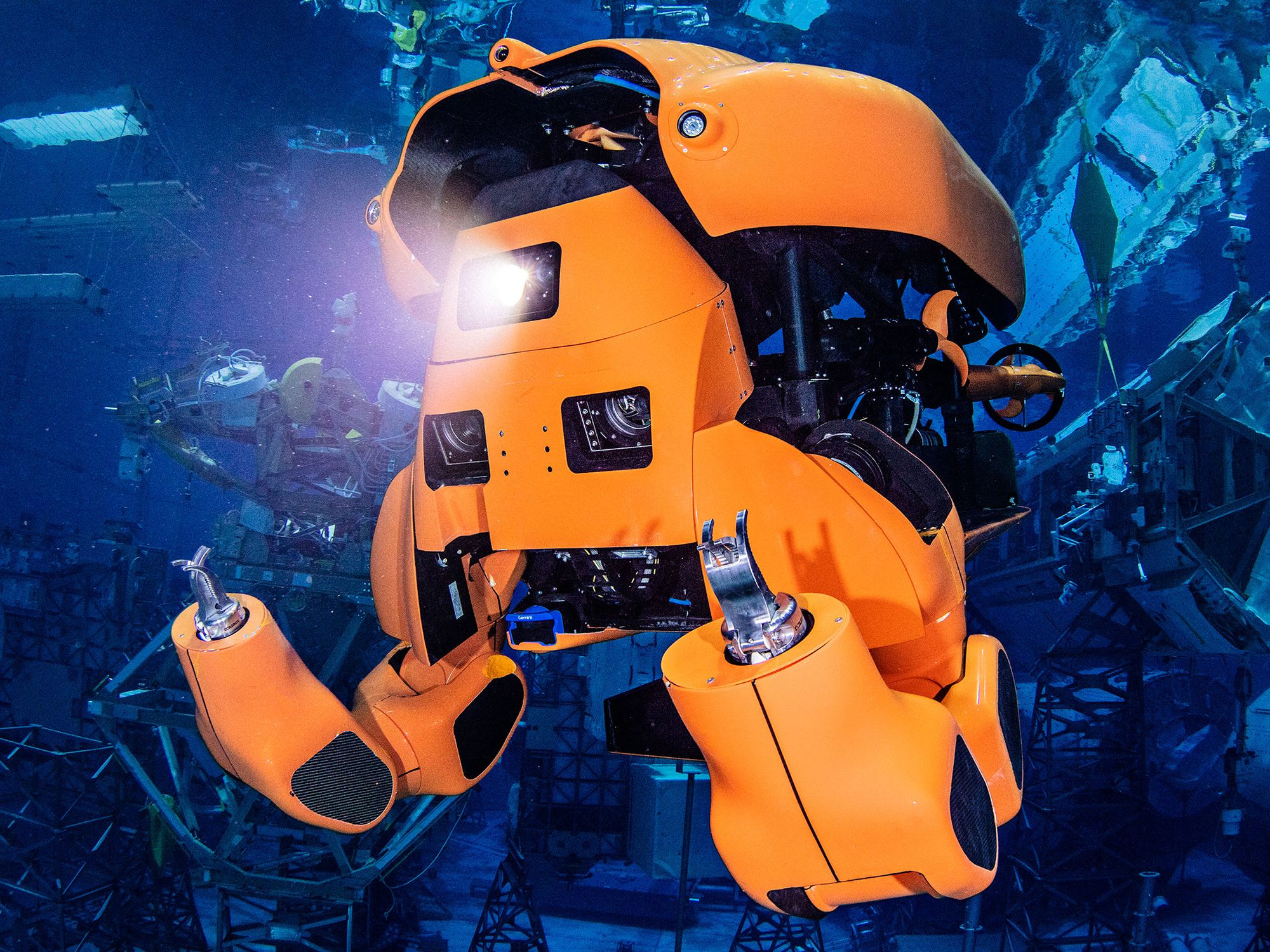 An orange half humanoid submarine robot with gripper hands shines a light underwater