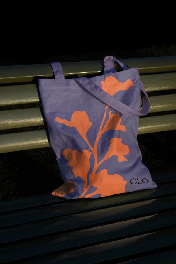On a green bench, a purple tote bag with an orange flower silhouette graphic and the word "CLO" in the bottom corner