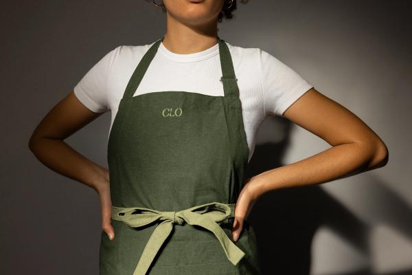 A woman stands with her hands on her hips wearing an apron with the word "CLO" embroidered on the front