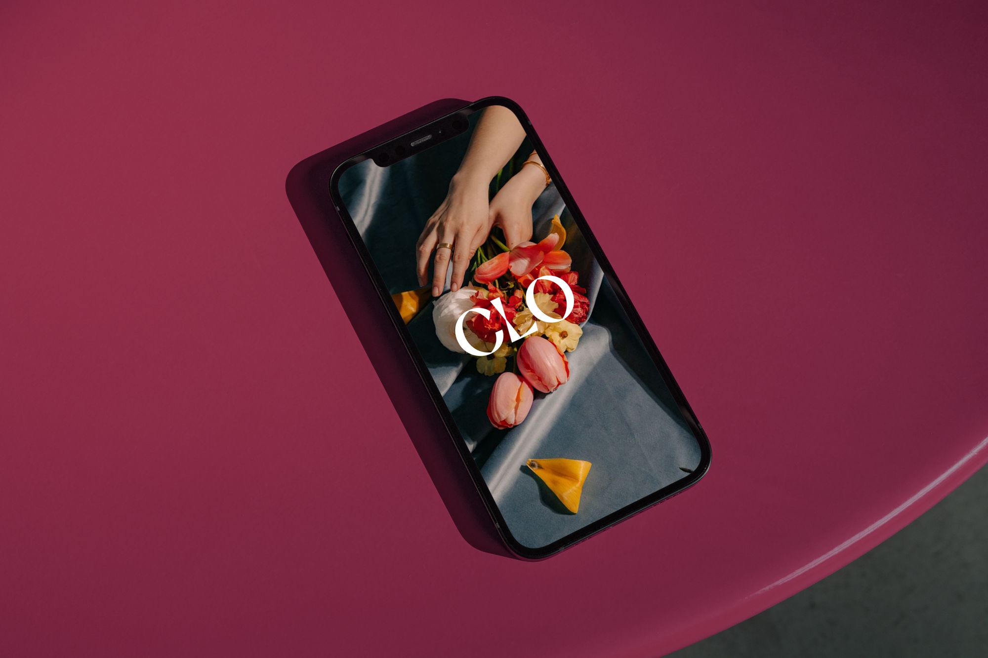 Smartphone on a pink table displays a photo of hands arranging tulips and roses with the letters "CLO" on the screen.