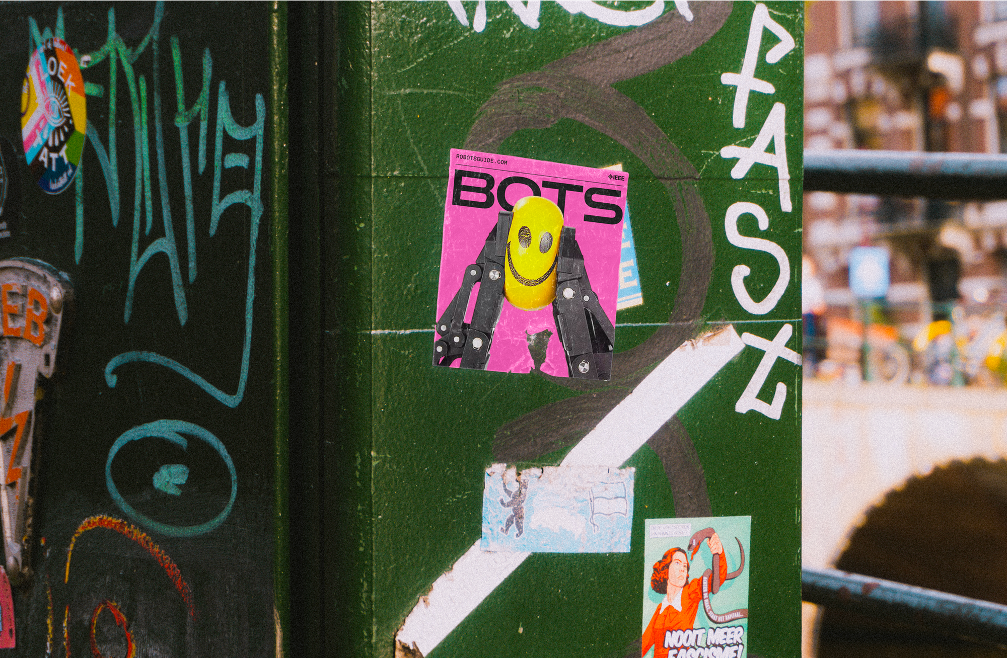 A green junction box in a public space is shown covered in graffiti and stickers, including one with a shortened BOTS logo showing a gripper robot